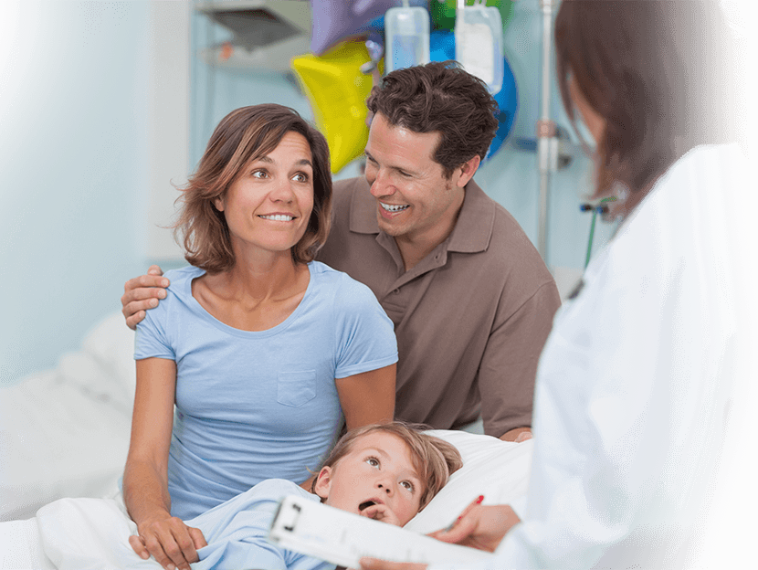 A woman and man standing next to a young boy in hospital bed.